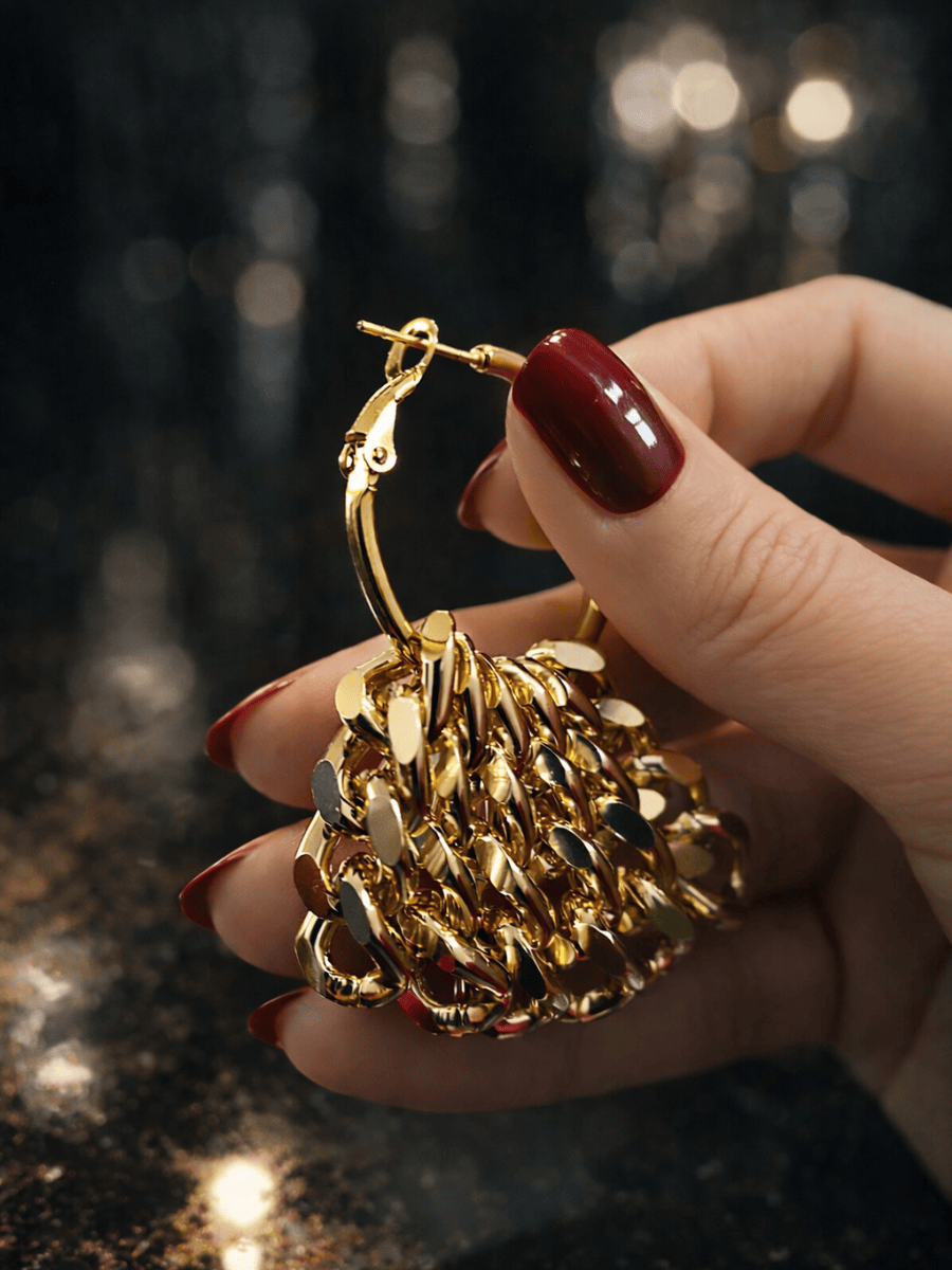 Gold chain hoop earring held by a hand with red nail polish on a dark background