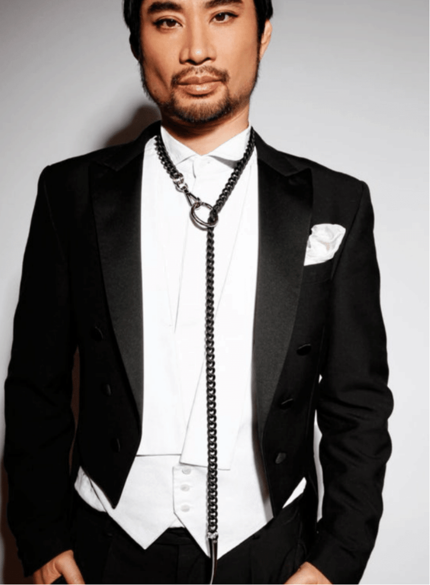 Man wearing a black tuxedo with a white shirt and a long black necklace against a gray background